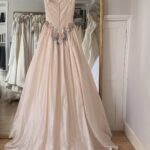 A pale pink wedding gown with floral appliqués hanging in front of a gold mirror.