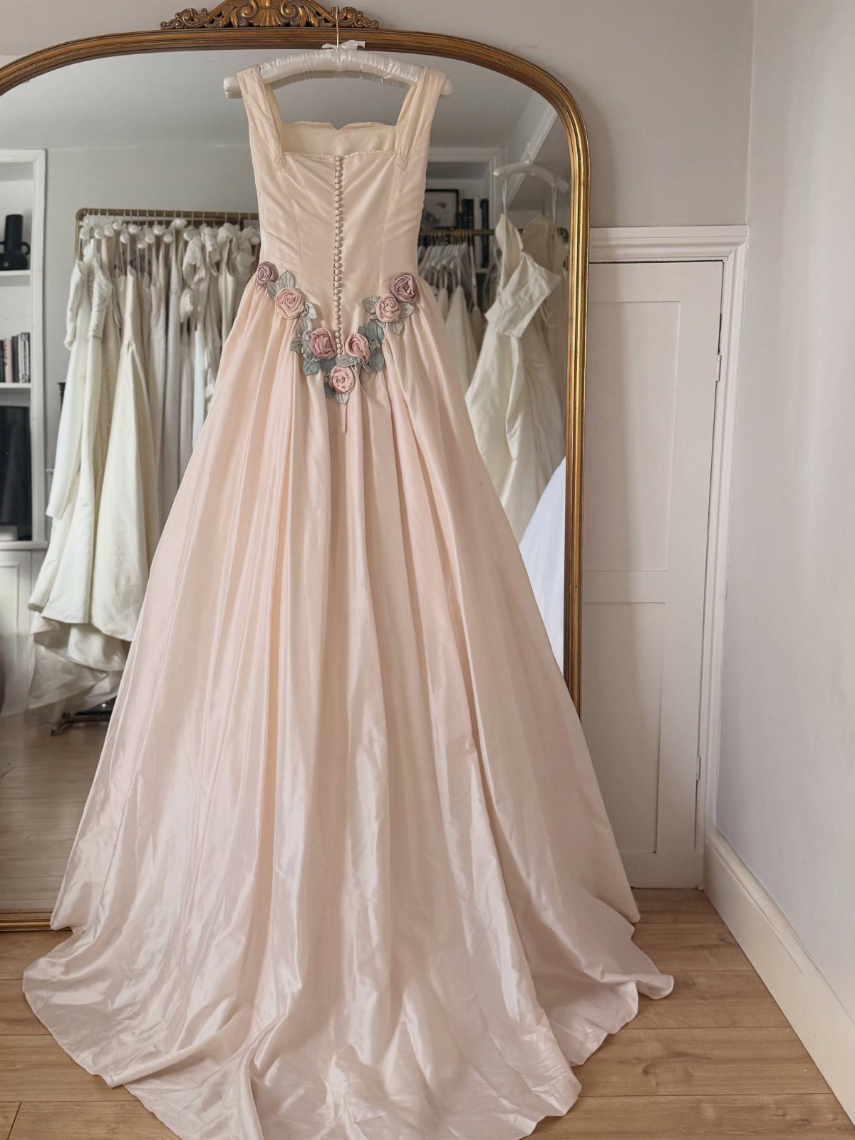 A pale pink wedding gown with floral appliqués hanging in front of a gold mirror.