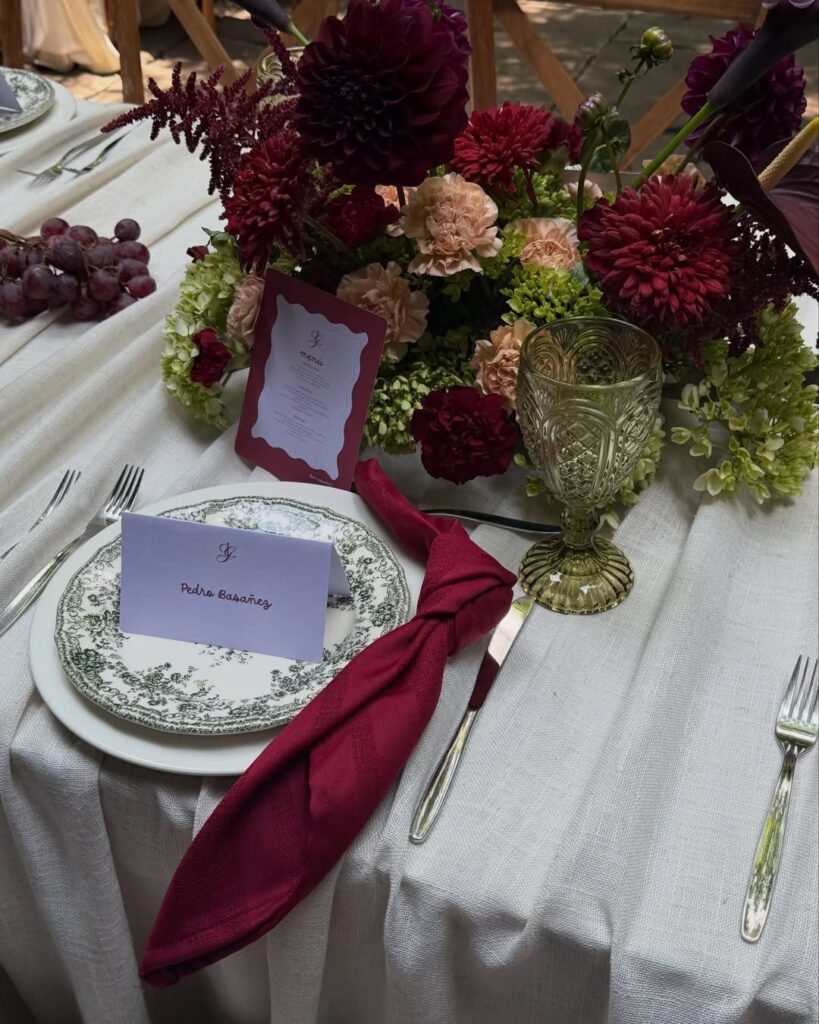 A wedding table setting with dark red floral arrangements, burgundy napkins, and gold glassware on white linen.