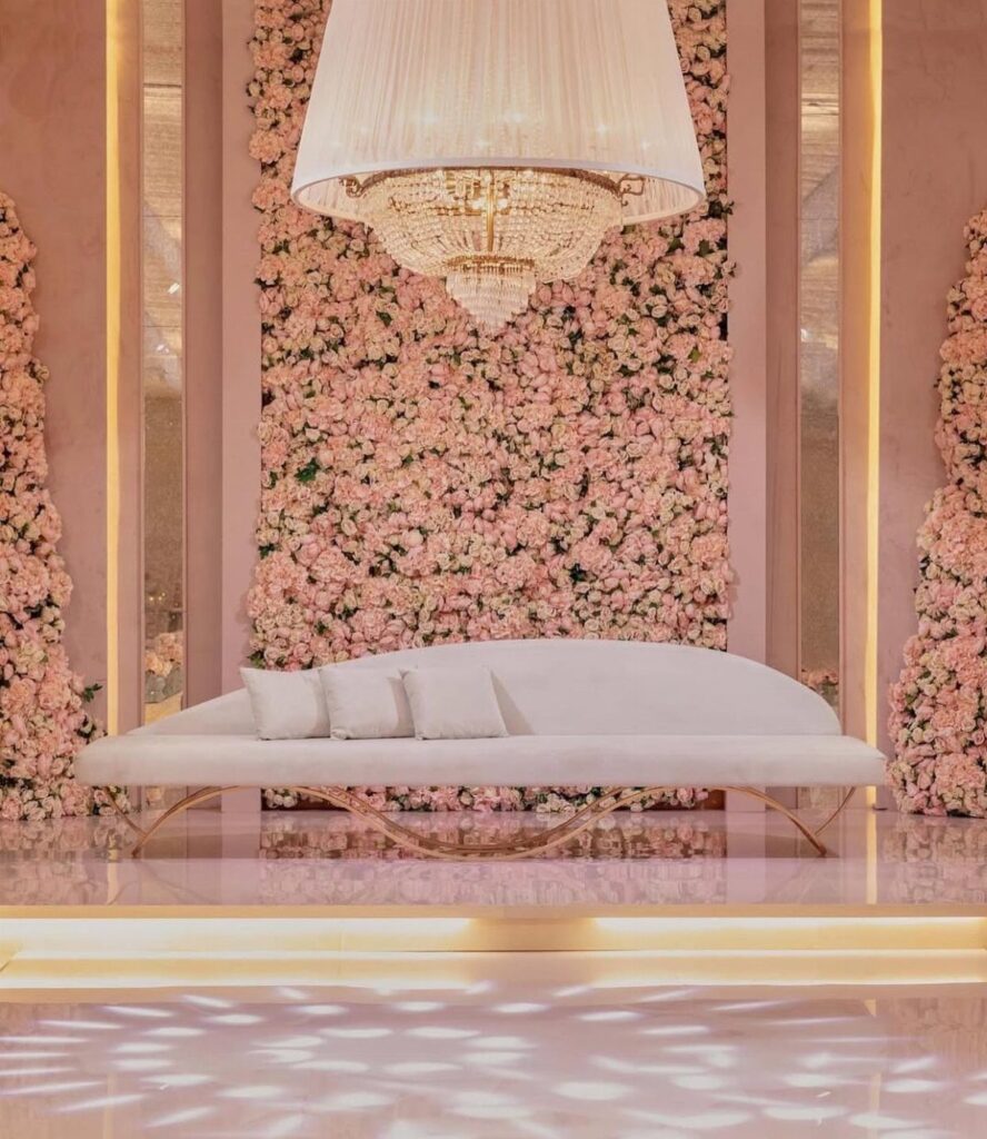 White curved sofa positioned in front of a large pink rose floral wall and crystal chandelier.