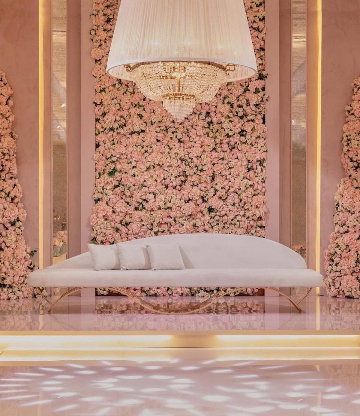 White curved sofa positioned in front of a large pink rose floral wall and crystal chandelier.