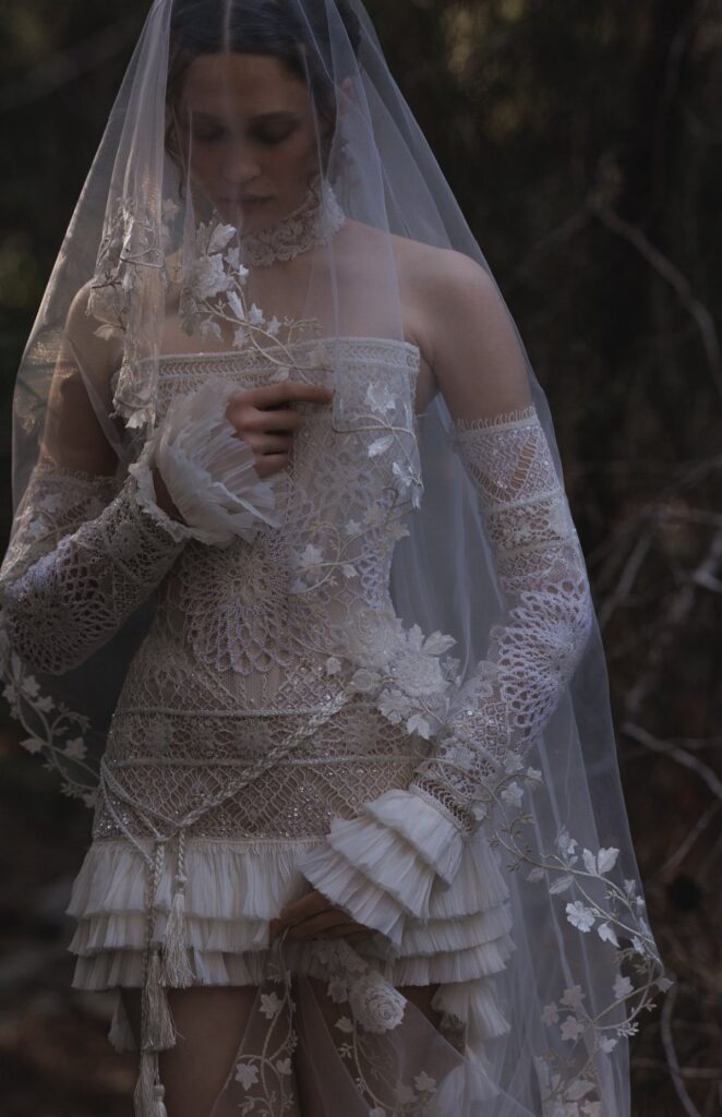 Woman in a short, embellished lace wedding dress and veil standing in a dark forest.