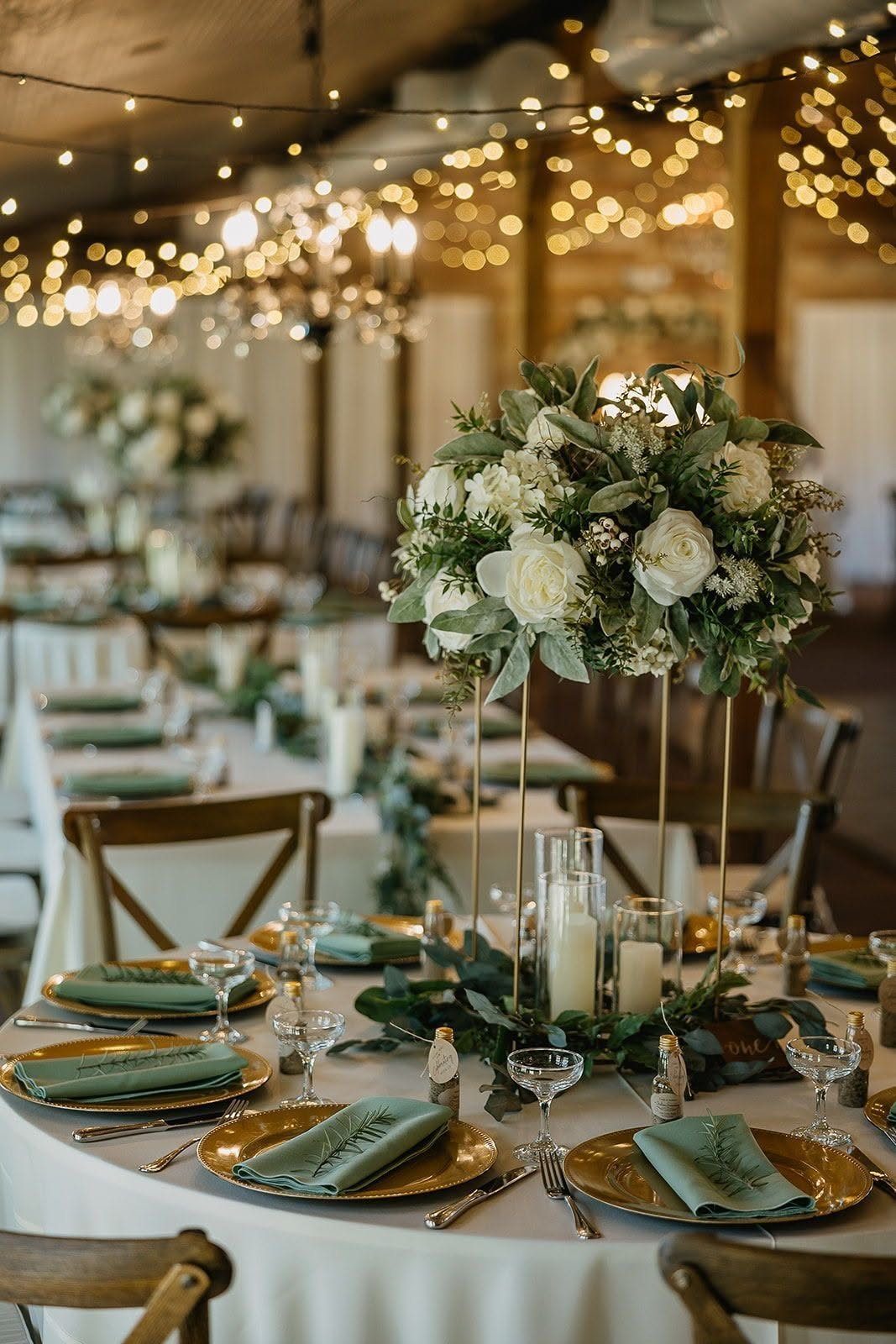 Wedding table setting featuring gold chargers, green napkins, and a tall white floral centerpiece with greenery.