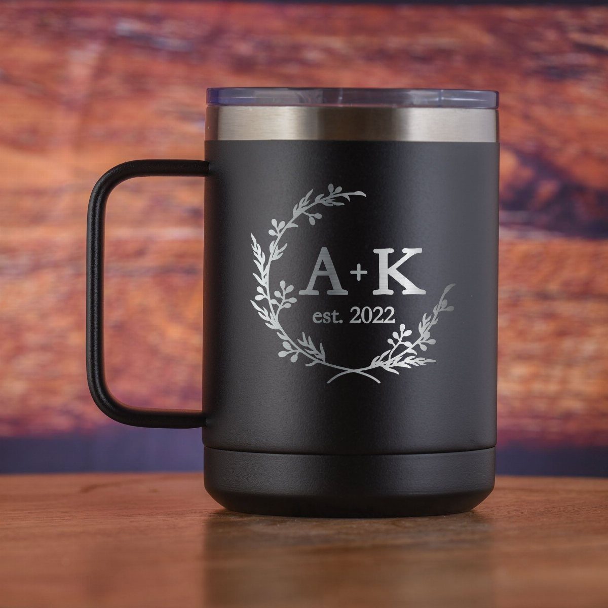 A matte black engraved travel mug with a floral wreath and initials on a wooden table.