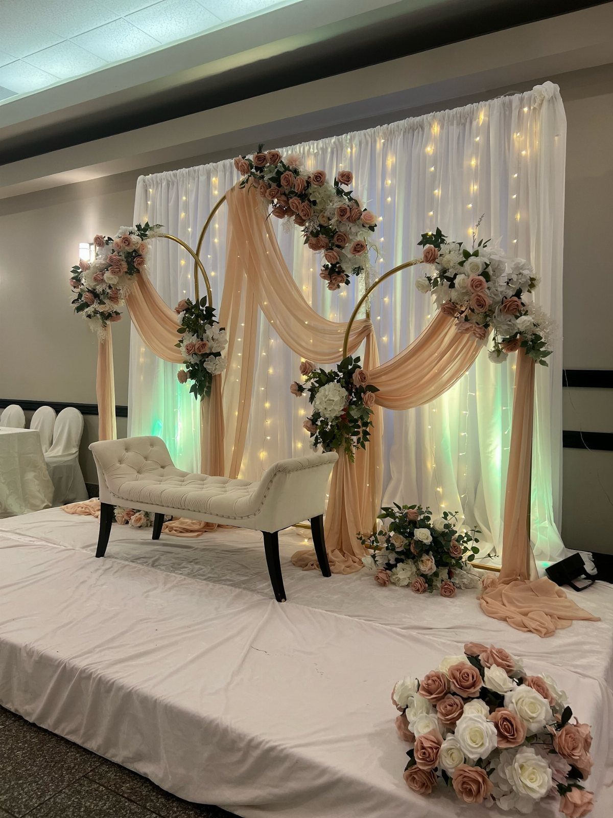 Gold wedding arch with peach drapes and floral arrangements in front of lit curtains.
