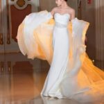 Woman in a white strapless wedding dress with a large, flowing, voluminous train walking indoors.