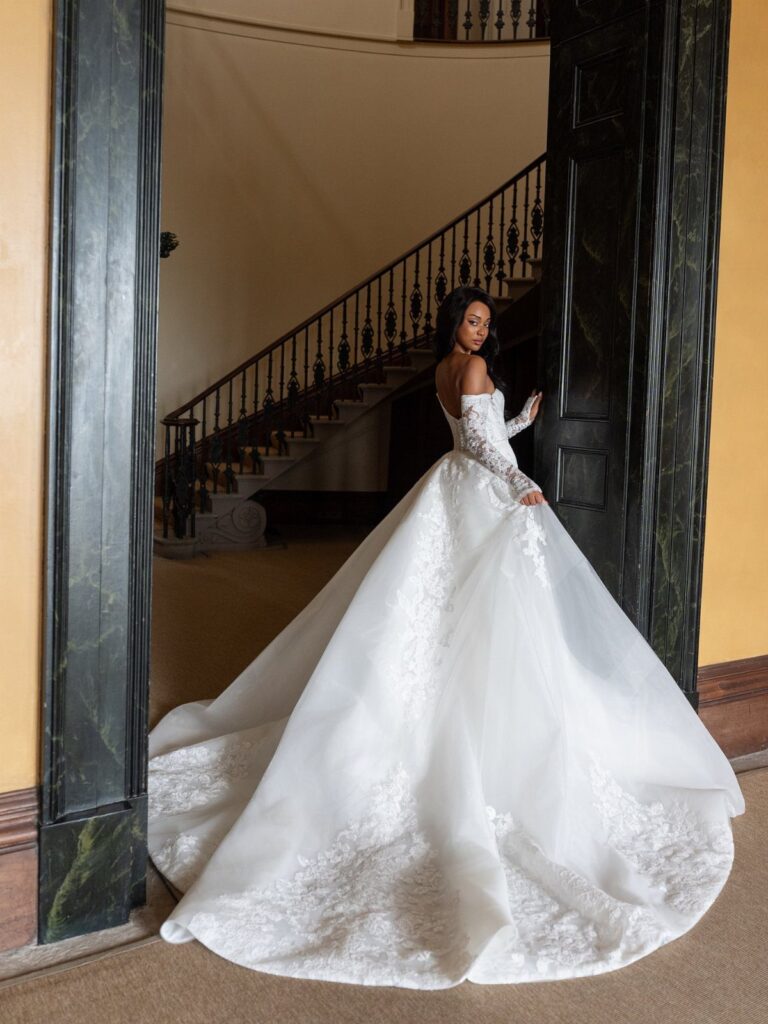 Woman wearing a white lace wedding dress with a large, voluminous overskirt standing in a doorway.
