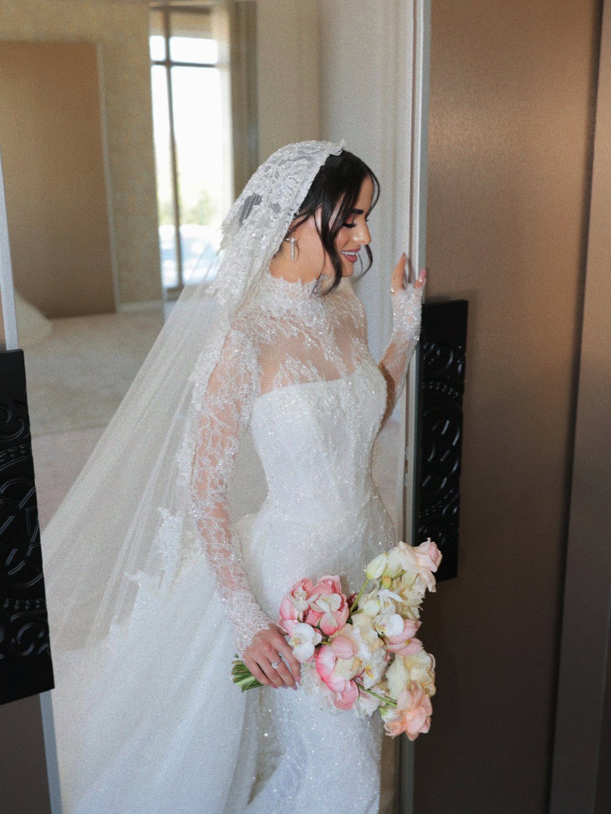A bride wearing a sparkling long-sleeved wedding dress and lace veil holding a floral bouquet.