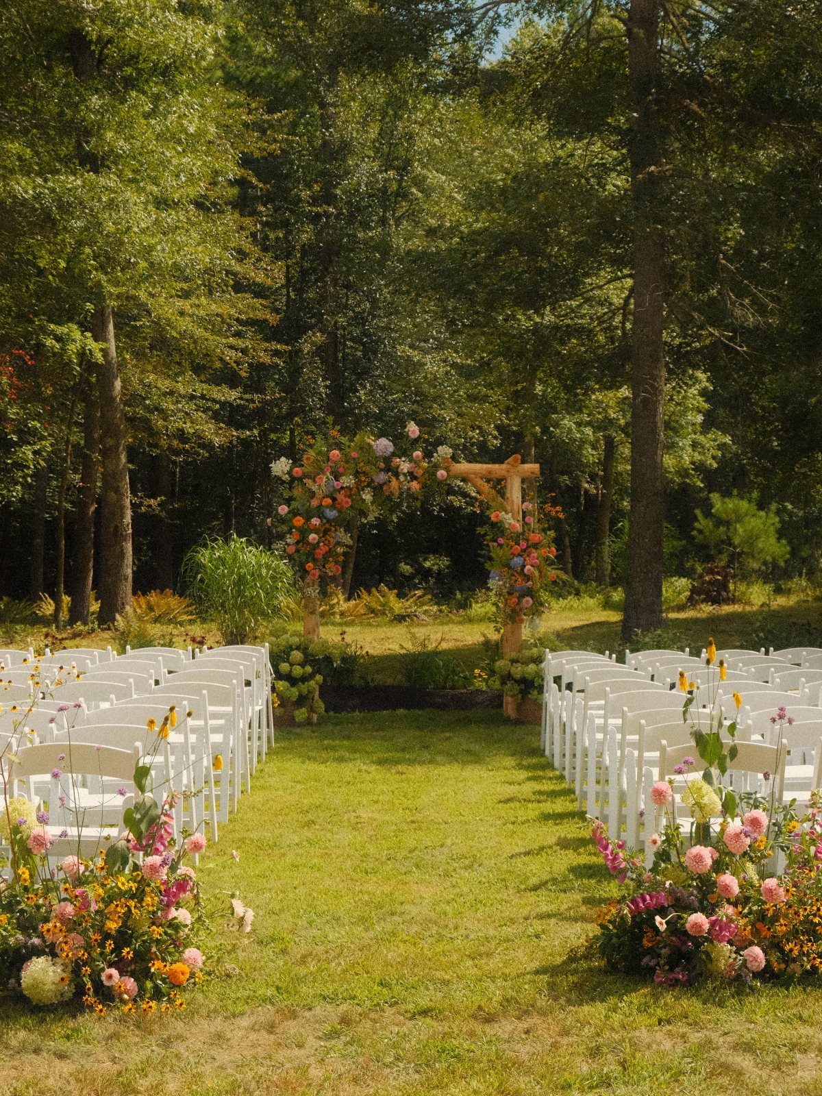 White wedding chairs arranged on grass facing a floral wooden arch in a forest.