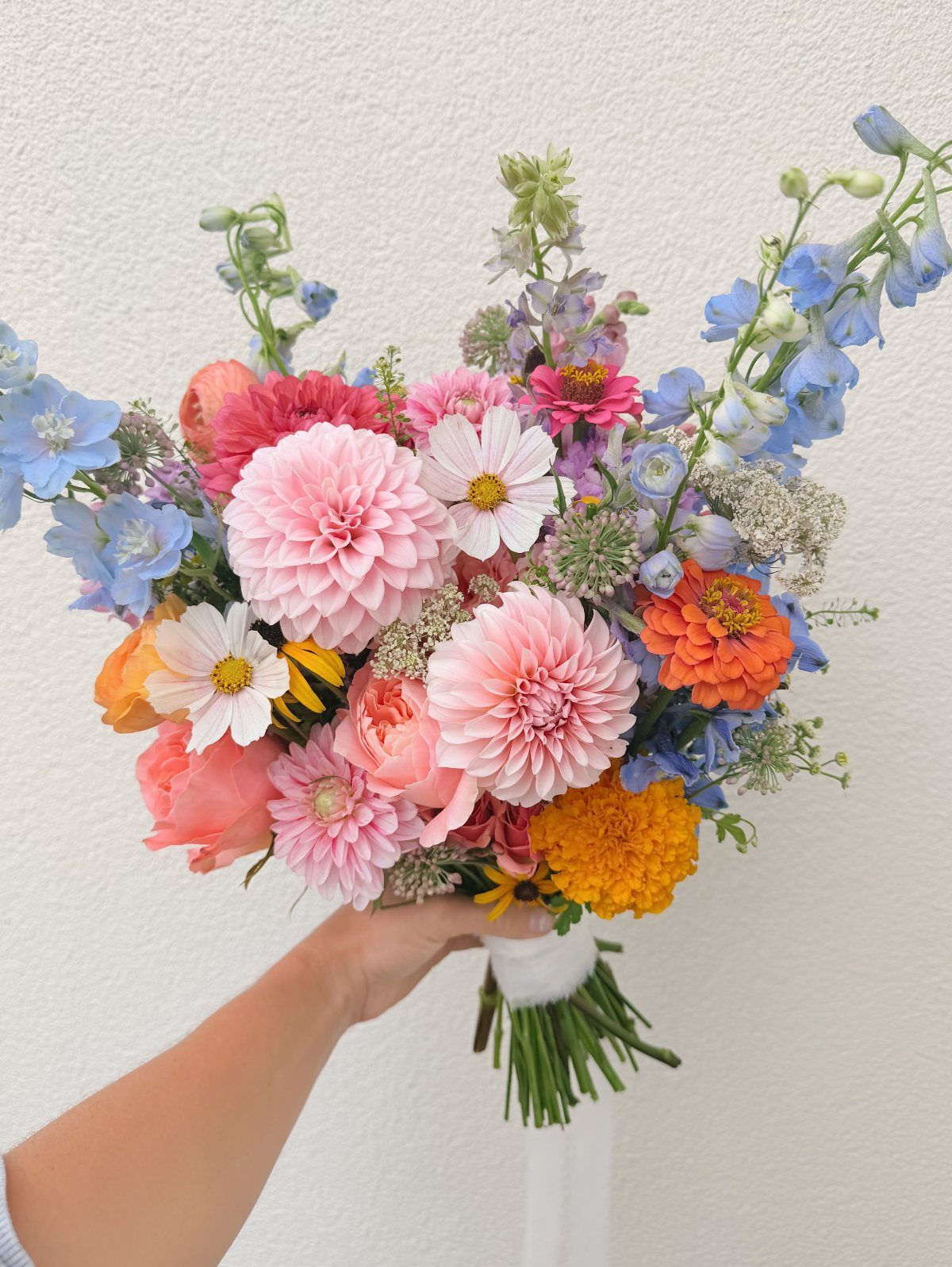 A hand holding a colorful wildflower bouquet with pink dahlias, orange zinnias, and blue flowers.